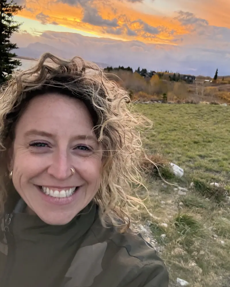 Dani Eve smiling outdoors with windblown curly hair, a grassy field and an orange sunset sky behind her.